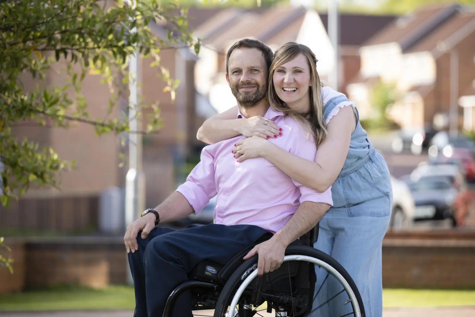 A smiling man in a wheelchair with a woman leaning in to hug him from behind, outside a row of houses