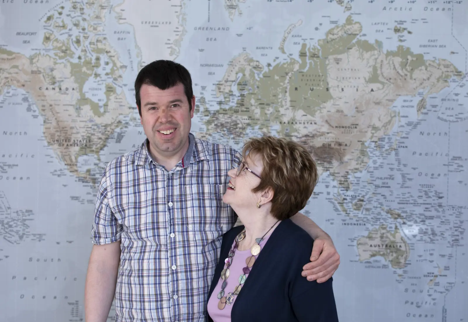 A smiling man with his arm around an older woman, who is laughing up at him, in front of a world map